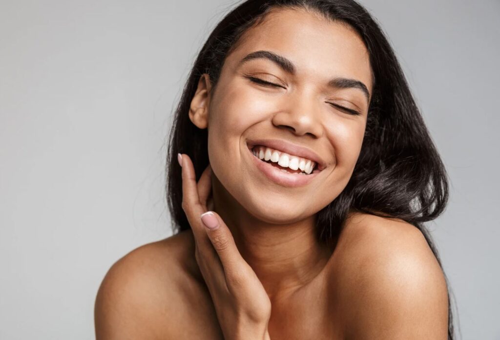 Smiling young woman with long dark hair, eyes closed and hand gently touching her neck, expressing joy and confidence. The background is a soft gray, enhancing her radiant and natural beauty. This image conveys a sense of happiness and self-assurance, ideal for beauty, skincare, or wellness content.